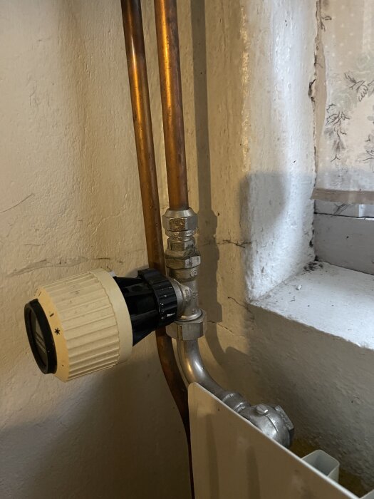 Radiator i tvättstuga med kopparrör och vit termostatventil monterad på väggen nära ett fönster.
