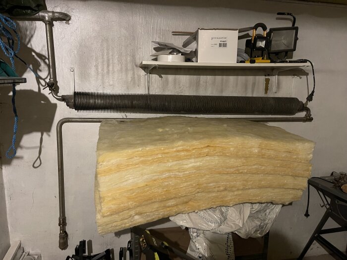 Gammalt rörelement i källare med synliga isoleringsmaterial och ventiler. På hyllan ligger verktyg och utrustning.