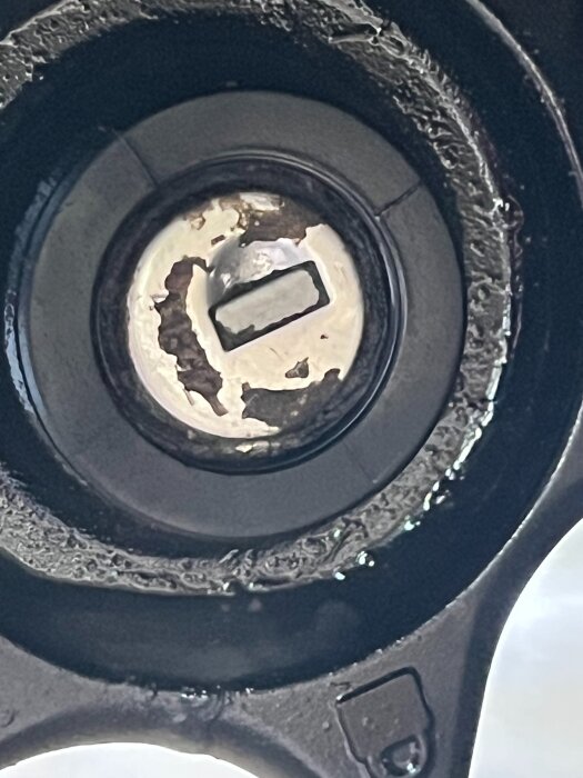Nära bild av en rund metallisk bilkomponent med en insatsfäste, omgiven av slitage och smuts.