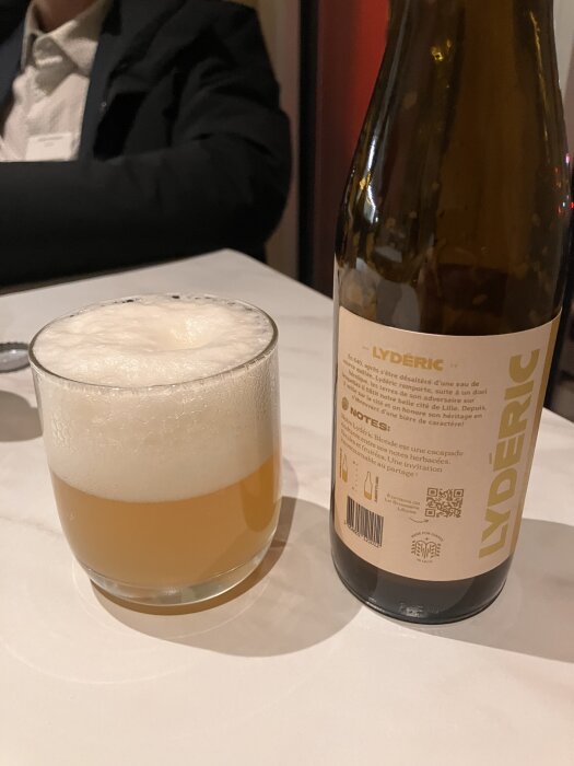 Glas med skummande öl bredvid en brun flaska med etiketten “LYDÉRIC” på ett bord. En person sitter i bakgrunden.
