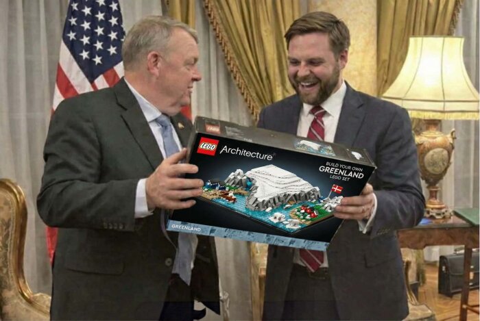 Två män i kostym ler och håller i en förpackning med LEGO Architecture-set "Build Your Own Greenland" i ett rum med amerikansk flagga i bakgrunden.