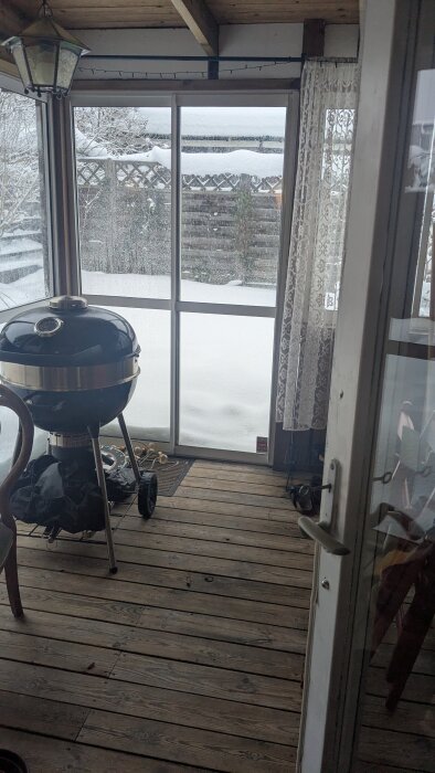 Uterum med trägolv, snötäckt utsikt genom glasdörrar med en vedeldad grill och gardin på sidan. Vinterväder utanför.