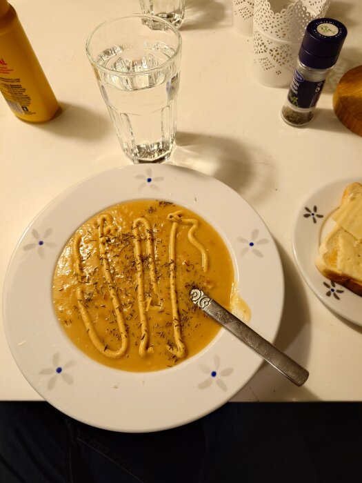 Krämig soppa i tallrik med ringlad sås ovanpå, bredvid står ett glas vatten, kryddburk och smörgås på tallrik.