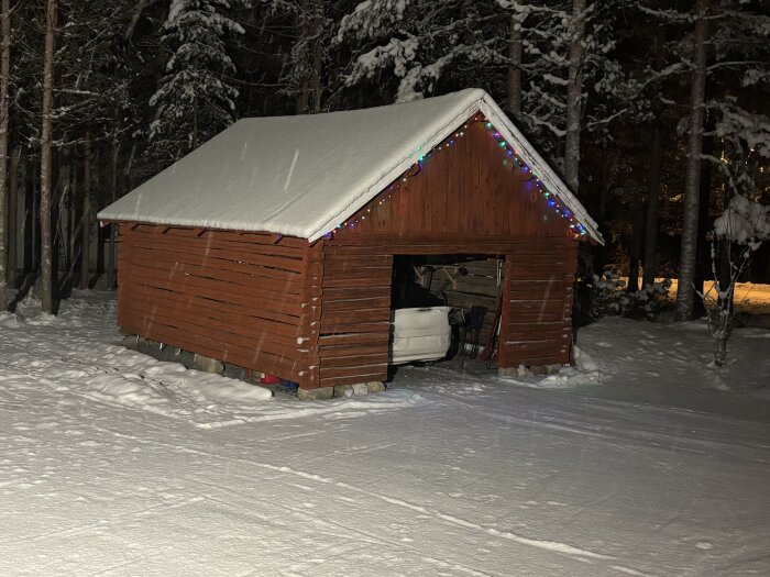 Rött träskjul i vintersnö med kulört julbelysning, omgiven av snötäckta träd.