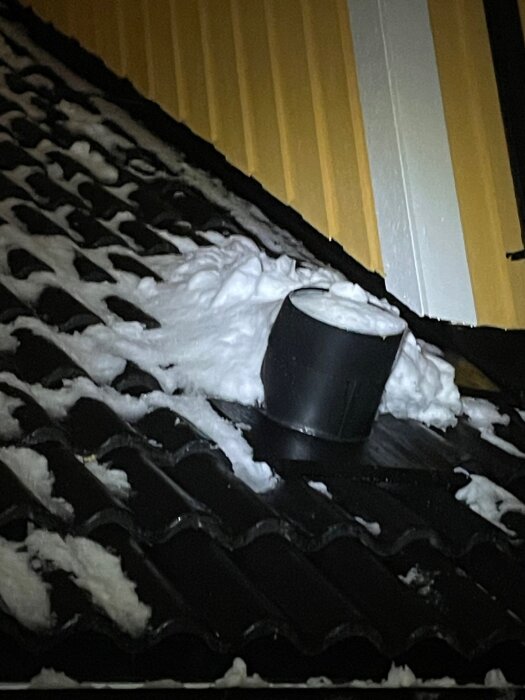 Tak med snö och svart takhuv, snön ligger på takpannor nära en gul vägg.