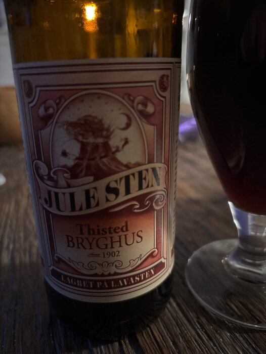 Brun flaska med etikett från Thisted Bryghus, text "Jule Sten", på ett bord bredvid ett glas med mörk dryck.