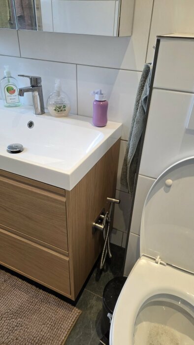 Badrumsinteriör med en bidédusch installerad vid ett träskåp bredvid toalett, sänka med tvålpumpar och spegelskåp ovanför.