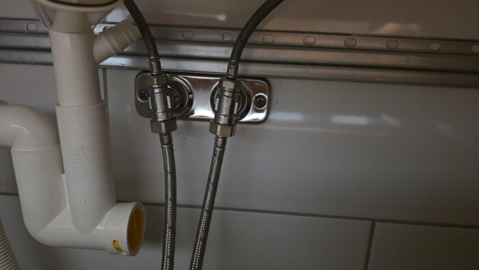 Två flexibla metallslangar kopplade till en väggmonterad anslutning under ett handfat, del av en bidéduschinstallation.