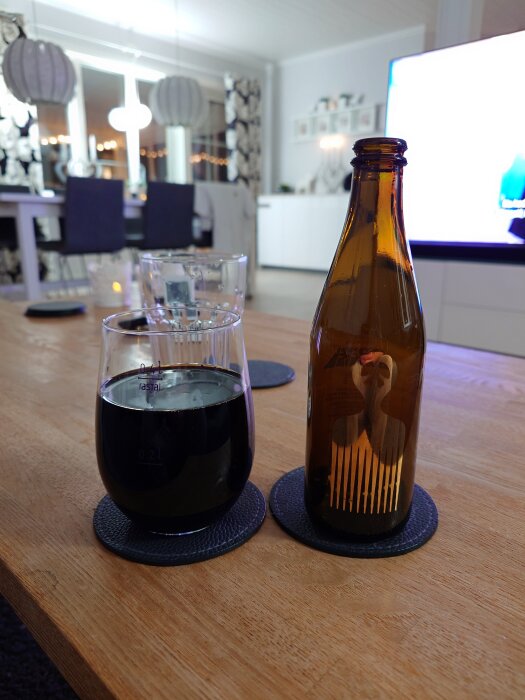 Flaska och glas på ett träbord, i bakgrunden en inomhusmiljö med bord och stolar.