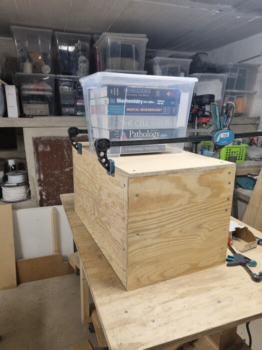En plywoodlåda under arbete med böcker i en plastlåda ovanpå, omgiven av olika verktyg och material i en verkstadsmiljö.