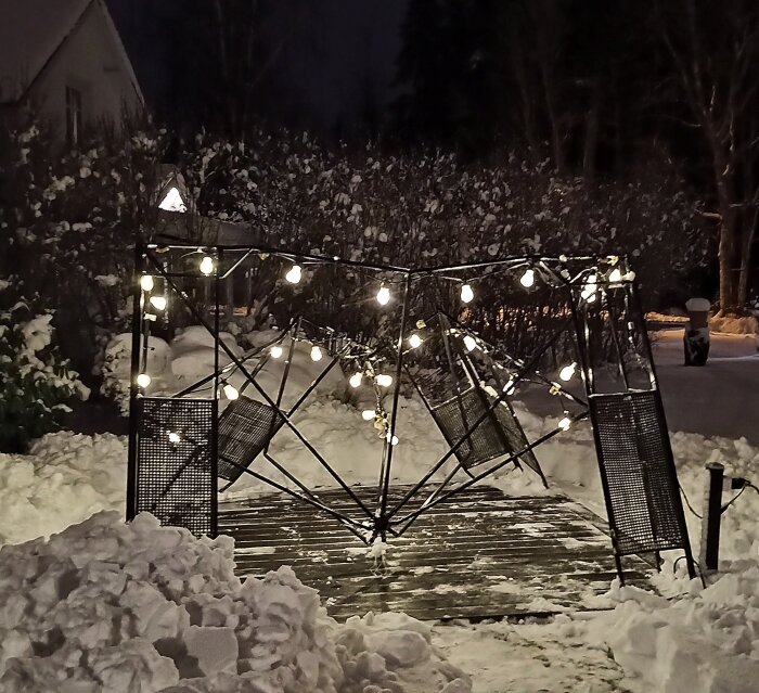 Nedsjunken paviljong i en snötäckt trädgård, dekorerad med ljusslingor, mot en bakgrund av buskar och träd.