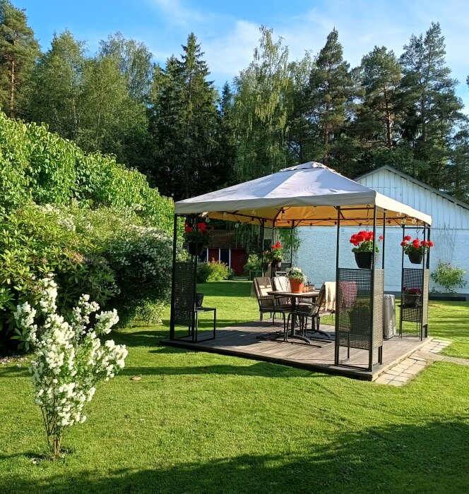 Paviljong i en grönskande trädgård, omgivet av blommor och träd. Enkelt bord och stolar står under taket, solig dag med blå himmel.