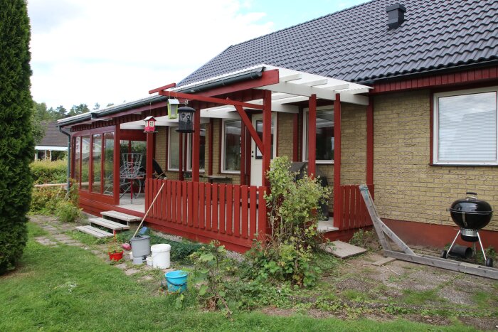 Röd veranda med lutande tak mot tegelhus, dekor och växter på gården, trädgårdsmöbler och utegrill på verandan.