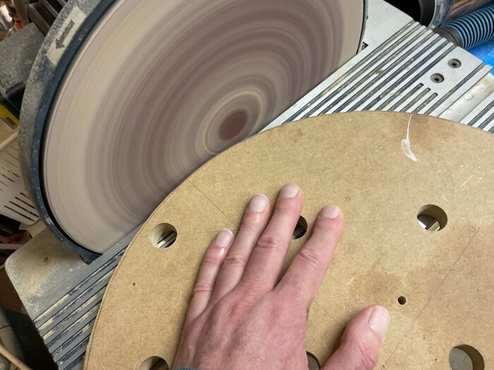 En hand håller en rund bit MDF med hål, slipas mot en roterande diskslipmaskin.