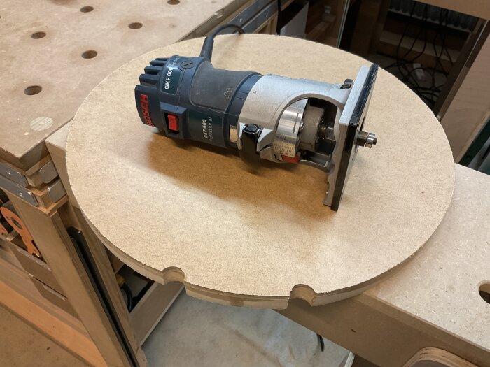 Rund MDF-fiberboardskiva med fräsverktyg ovanpå, verkstadsmiljö i bakgrunden och halvcirkelformade urtag längs kanten.