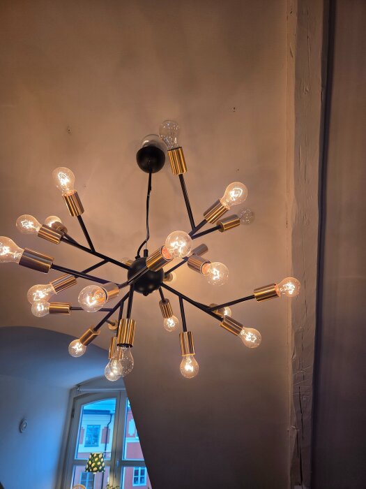 Modern taklampa med flera glödlampor i olika riktningar, monterad i ett rum med snedtak och ett fönster som släpper in dagsljus.