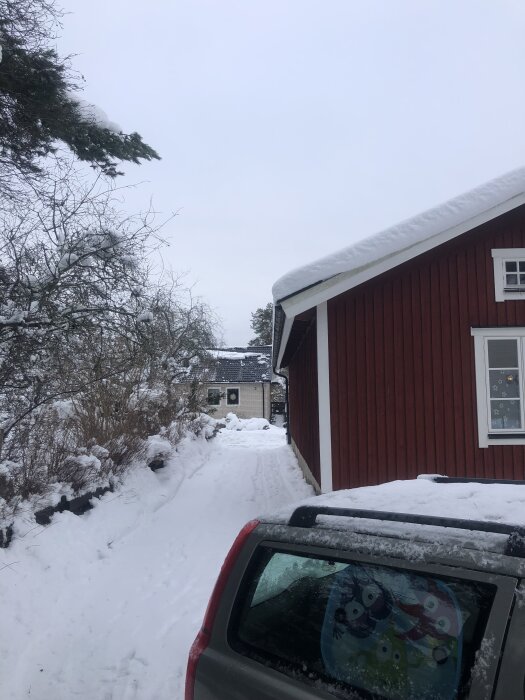 Smal uppfart med snö, omgiven av häck och rött hus, bil syns delvis i förgrunden, grå himmel och snöiga tak.
