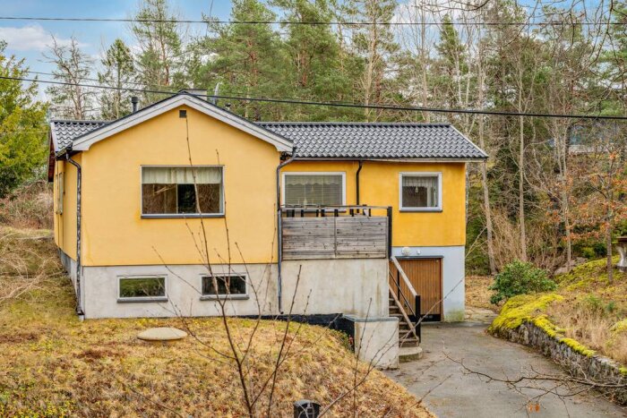 Ett gulmålat hus med grå takpannor, sliten altan och trappuppgång på framsidan, omgärdad av träd och buskage.