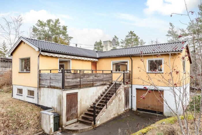 Gult hus med svart tak, träaltan och trappa på framsidan. Framsidan visas med ingång, trädgård och garage i fokus.