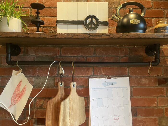 Hylla på tegelvägg med kittel, dekoration, växt och ljusstake ovanpå; nedanför hänger ritning, skärbrädor och kalender från en stång.