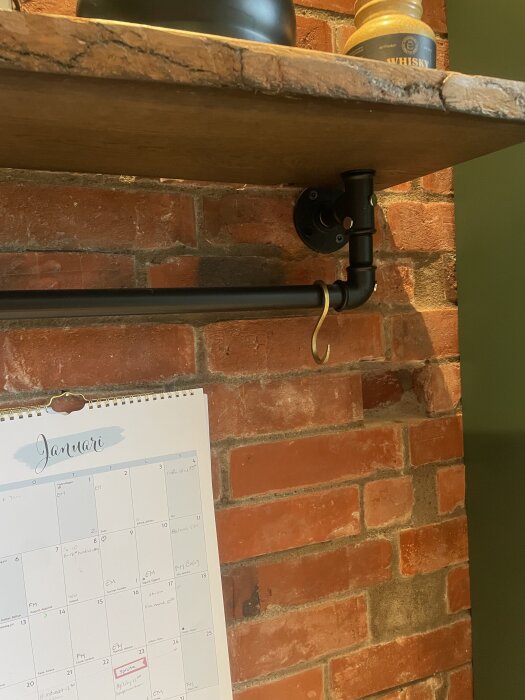 Hylla av trä och metall med whiskyburk ovanpå, kalender för januari hänger på röd tegelvägg.