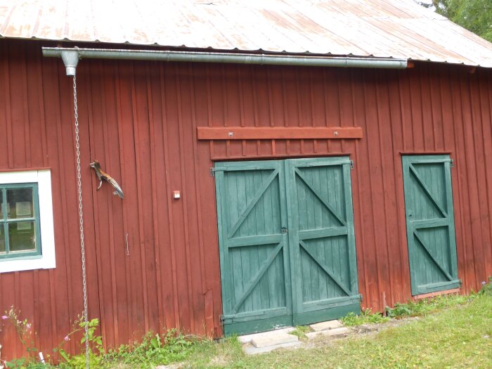 Röd träbyggnad med gröna dörrar och ett fönster, takränna och regnkedja, samt en rävsvans på väggen.