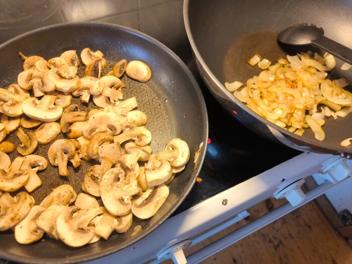 Stekta champinjoner i en stekpanna bredvid stekt lök i en wokpanna på en spis.