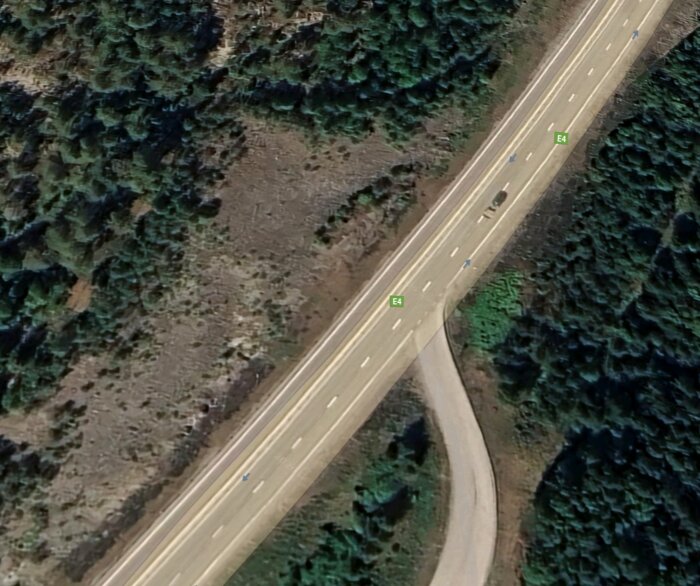 Flygbild av E4-väg nära Höga Kusten, omgiven av skog och en mindre anslutningsväg, sedd från ovan.