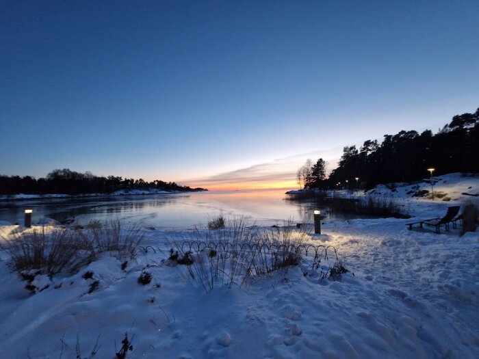 Snötäckt kustlandskap i skymningen med belysning längs strandpromenaden och solnedgång i bakgrunden.