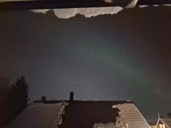 Nattbild av snötäckt tak och några träd under en stjärnklar himmel med svagt norrsken i bakgrunden.