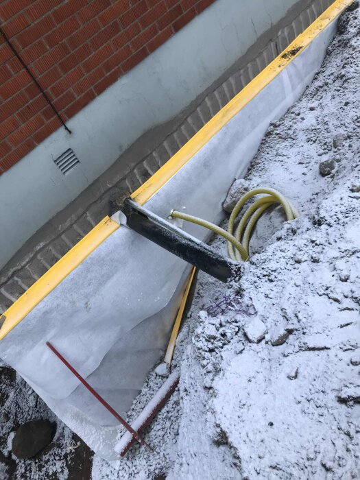 Dräneringsjobb vid husgrund med murad vägg, snö på marken, gul och röd dräneringsslang synlig, isolering och skyddsplast uppsatt mot väggen.
