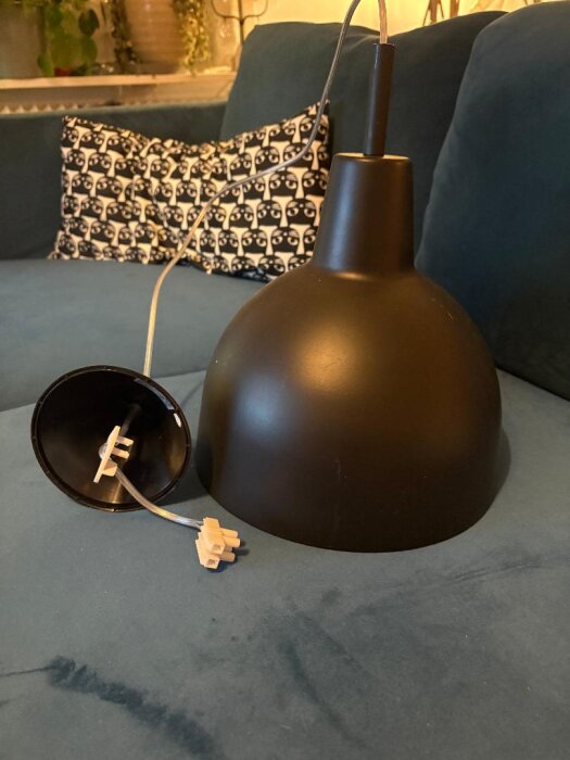 Svart taklampa med plastkopp och elektrisk anslutning på en blå soffa med kudde i bakgrunden.