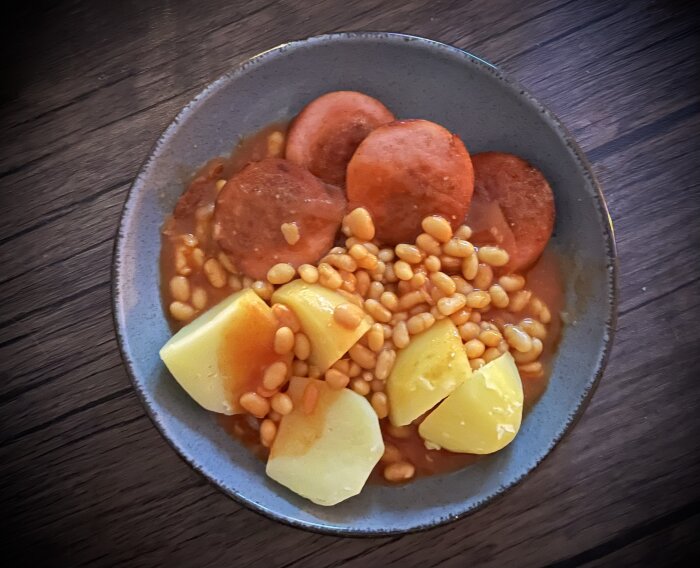 Tallrik med skivad korv, bönor i tomatsås och kokt potatis på ett bord.