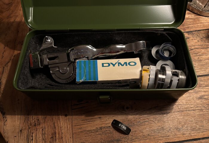 Dymo präglingsapparat i en grön låda med kassetter och en pappersförpackning, liggande på ett träbord.
