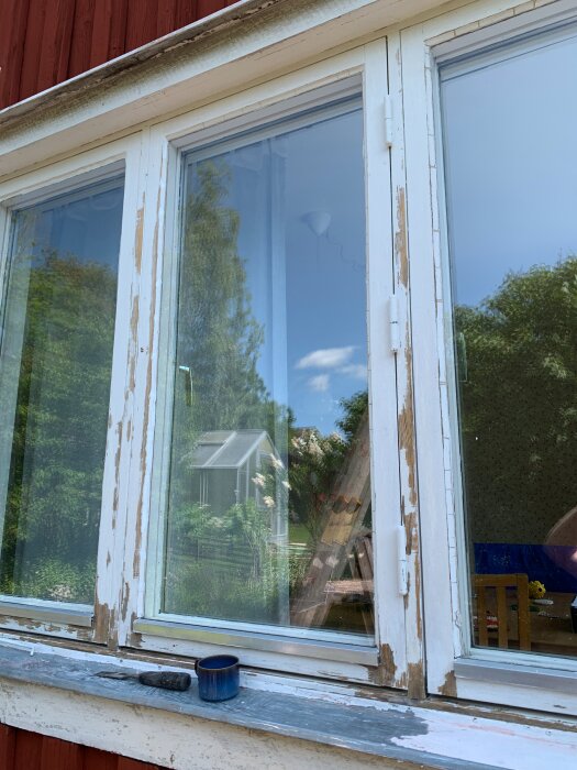 Slitet vitmålat fönster med skrapad färg på ett rött hus. På fönsterbläcket ligger en liten blå burk och pensel. Reflektioner av träd och trädgård.