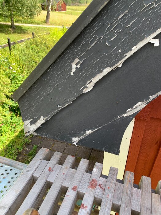 Flagnande svartmålad vindskiva på husgavel med röda väggar, visande skadat och avflagnat trä. Träställning i förgrunden, trädgård i bakgrunden.