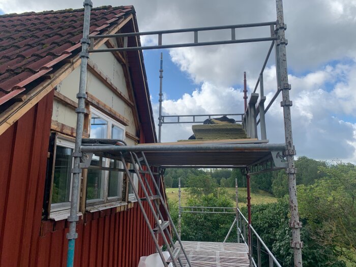 Byggställning vid röd husgavel med öppet träarbete runt fönster; takpannor synliga, himmel och träd i bakgrunden.
