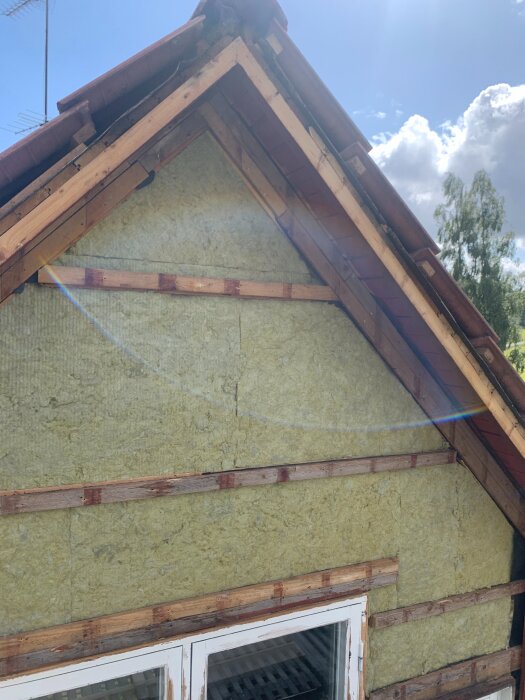 En gavel av ett hus under renovering, med exponerad isolering och delvis borttagen panel. En solfläck syns över bilden.