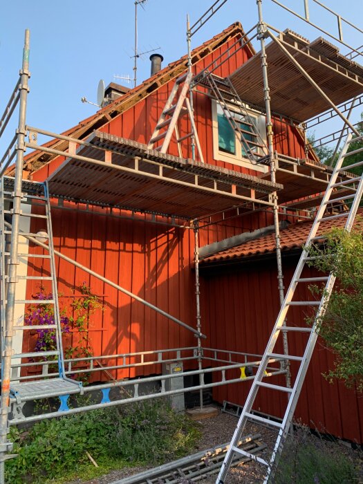 Rött hus med byggställning vid gaveln, stege lutad mot ställningen. Takplåtar och fönster synliga. Soligt väder, blå himmel.