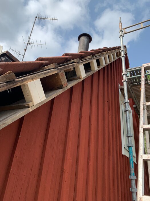 Röda träpaneler på ett hus, med synliga vindskivor under takpannor. Byggställning intill och antenner i bakgrunden mot en blå himmel.