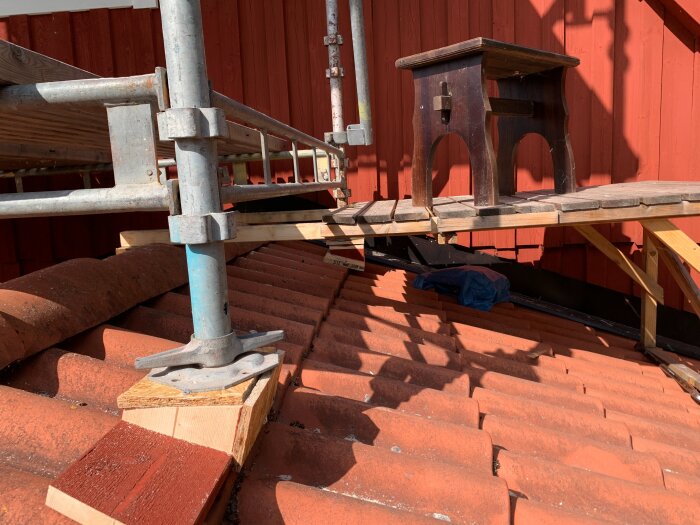 Byggställning och träplattform över takpannor vid ett hus med röda vindskivor, visar renoveringsarbete på taket.