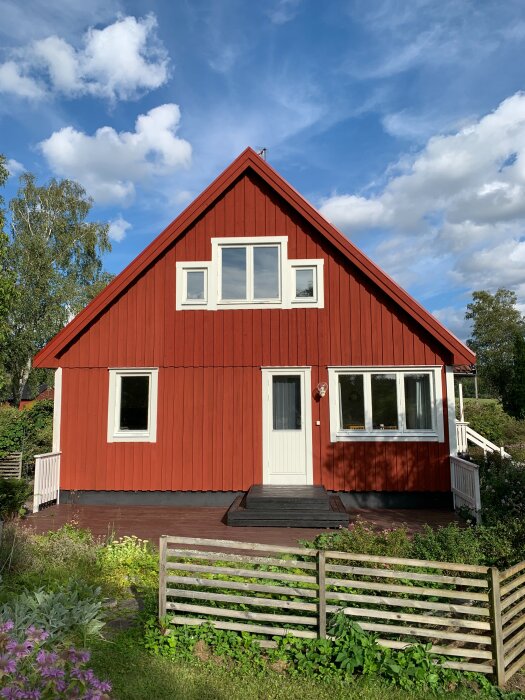 Rött 1,5-planshus med vita fönster och dörr, omgiven av grönska och träd, under en blå himmel med moln.