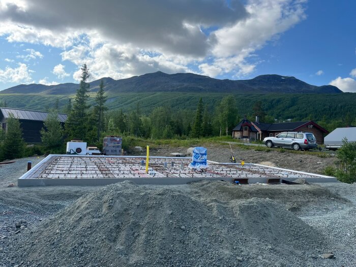 Grundläggning för fritidshus med fjäll i bakgrunden, grushög i förgrunden och en bil parkerad vid stugan.