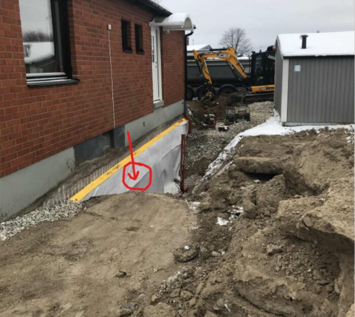 Urgrävning vid husets grund med markerad fuktgenomträngning; grus och jordhögar samt en synlig grävmaskin i bakgrunden.