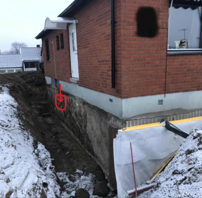 Urgrävning vid husgrund med pil som markerar fuktskadad yta; snö på marken. Tegelhus syns ovanför grunden och dränering är påbörjad.