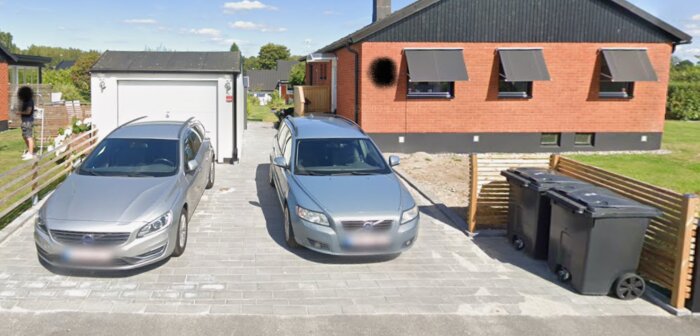Två bilar parkerade på en stenlagd garageuppfart framför ett rött tegelhus. Sopkärl står vid sidan av uppfarten.