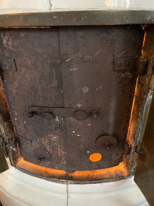 Närbild av en gammal braskamins dörr i metall med rost och ett runt ventilationshål. Dörren är delvis öppen och en orange glöd syns inuti.
