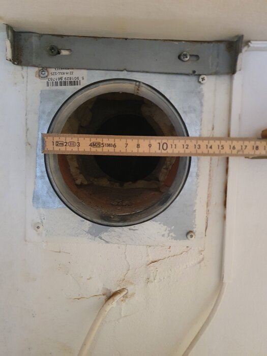 Hål för köksventilation med omgivande metallram och trälinjal som mäter cirka 110 mm diameter. Ingen synlig tätning runt kanten.