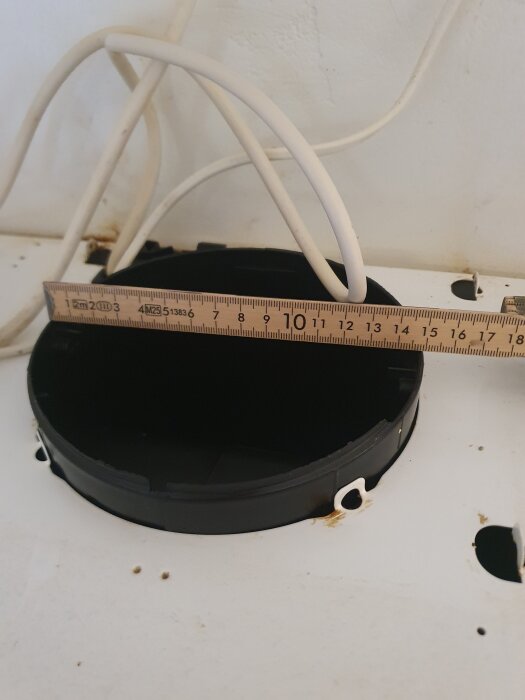 Öppning för köksventilation med linjal som mäter diameter, omgiven av kablar och ett svart rör, på vit yta.