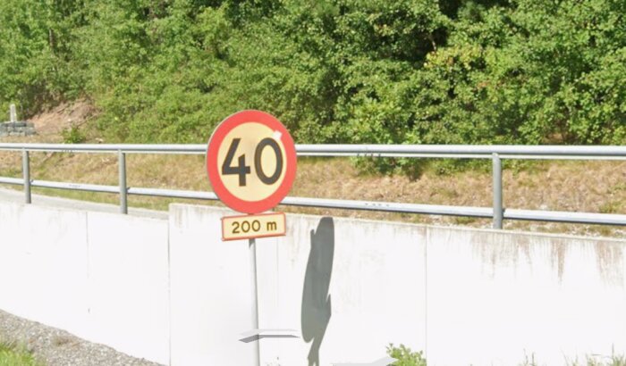 Vägskylt med hastighetsgräns 40 km/h, cirka 200 meter innan begränsningen börjar gälla, omgiven av vägräcke och grönska.